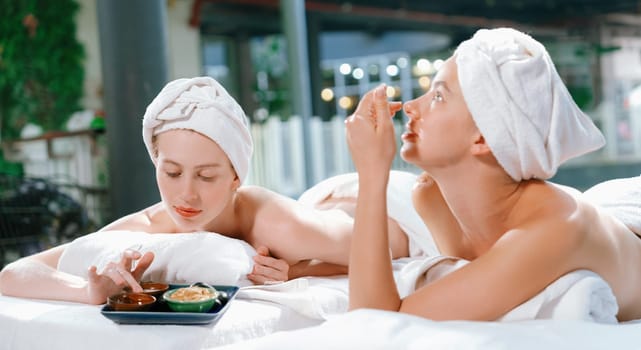 Couple of beautiful young girls lie on spa bed during interested in homemade beauty facial mask. Attractive woman touching herbal facial mask. Surrounded with nature environment. Tranquility.