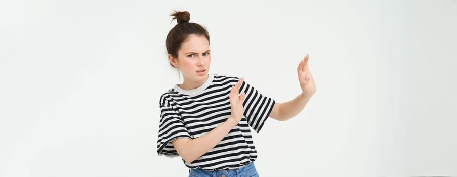 Image of woman expressing aversion, dislike something, looks apalled, extends hands forward in defensive gesture, rejecting, refusing, standing over white background.