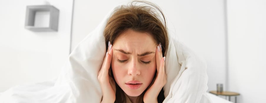 Close up of woman feeling unwell at home, lying in bed under white blankets and frowning, touching head, has headache, migraine, recovering from covid.