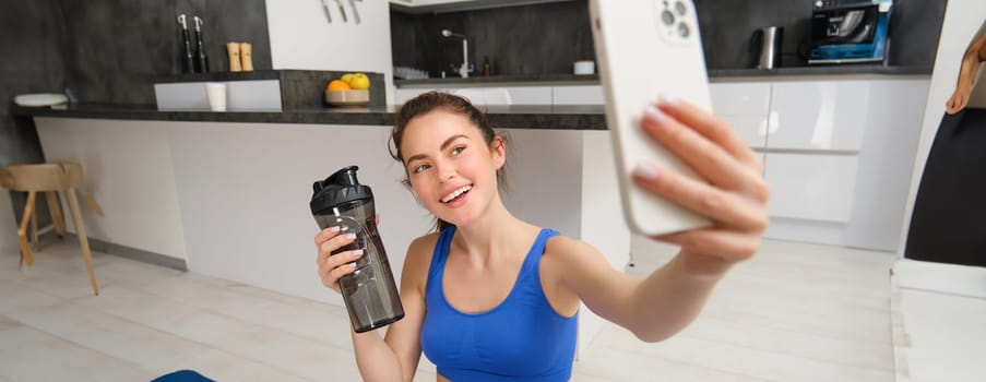 Portrait of sportswoman takes selfie with water bottle in living room, holds smartphone and poses for photo while doing fitness workout.