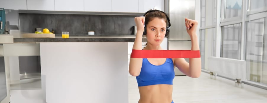 Home workout and wellbeing. Focused young fitness woman, stretching elastic resistance band with arms, doing muscle exercises in living room, listening music in wireless headphones and exercising.