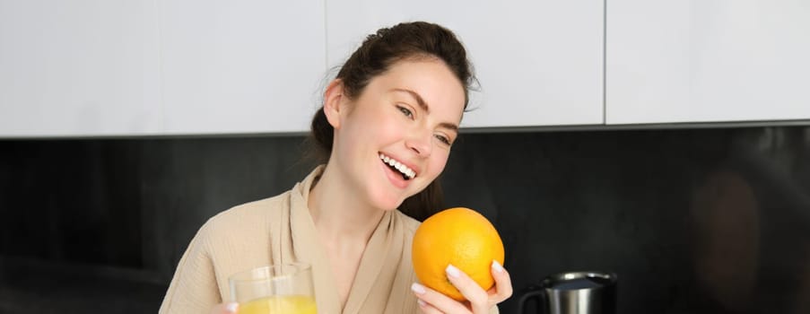 Portrait of attractive young modern woman, posing in kitchen, holding glass of juice and an orange, laughing and talking to someone aside.