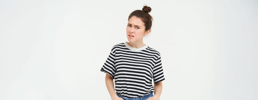 Portrait of confused young woman, looking with disbalief, raising her eyebrows and staring shocked at camera, standing over white background.