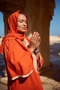 Portrait of beautiful serene Arabian Muslim woman in bright orange authentic dress and her head covered in hijab, praying with her eyes closed, hands up and palms together. People and religion concept