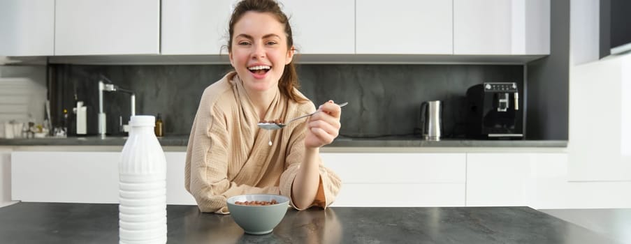 Portrait of beautiful young and healthy woman in bathrobe eats her breakfast in kitchen, has cereals with milk and smiling.