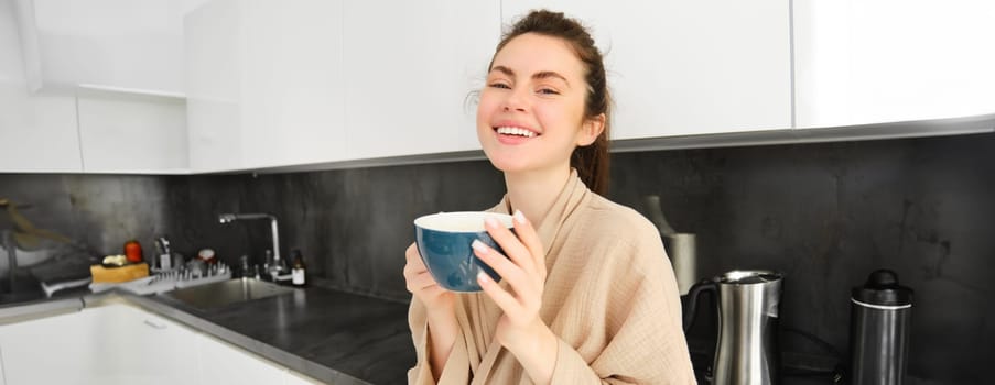 Beautiful young woman enjoys her morning mug of coffee, drinking tea from cup in the kitchen, wearing bathrobe and smiling at camera.