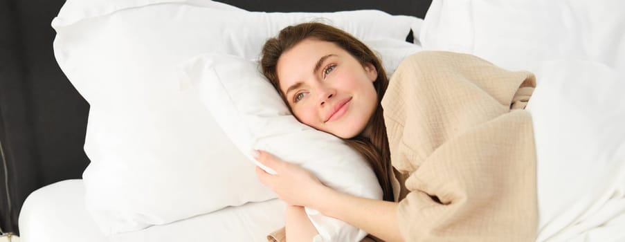 Gorgeous woman resting in bed wearing pyjamas, hugging her pillow and smiling.