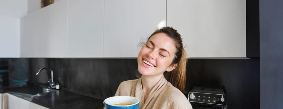 Portrait of carefree smiling woman drinking coffee, standing in kitchen with delighted, pleased face expression, waking up in the morning and having cappuccino.