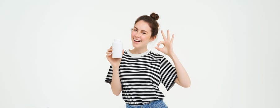 Image of smiling girl recommends dietary supplement, bottle with vitamin c, fish oil treatment, stands over white background.