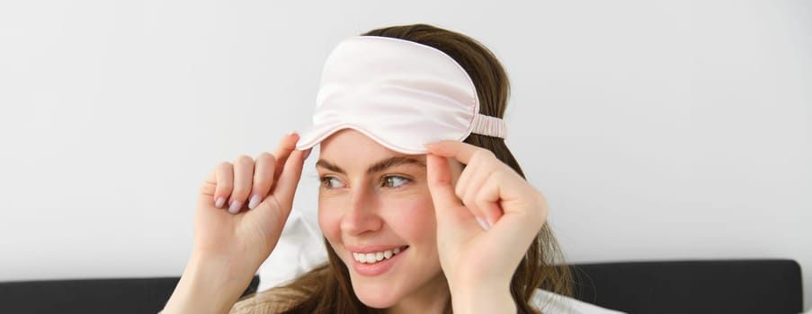 Portrait of smiling young modern woman open her eyes in morning, waking up and taking off sleeping mask, looking outside window, resting in bed, lying in bedroom.