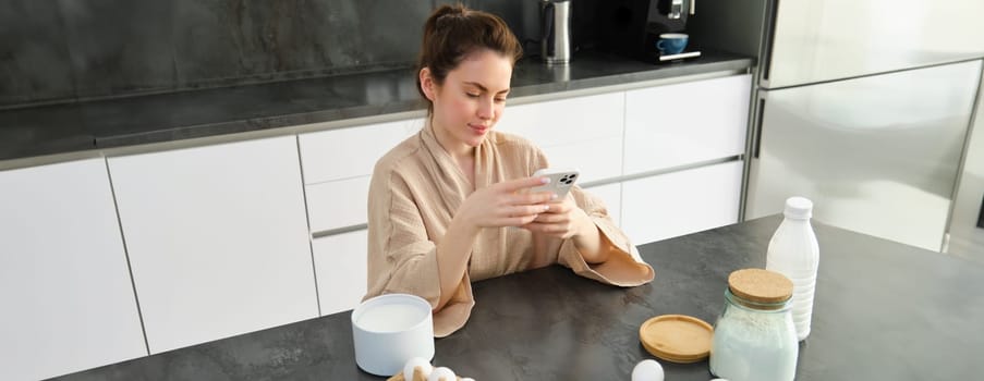 Attractive young cheerful girl baking at the kitchen, making dough, holding recipe book, having ideas.