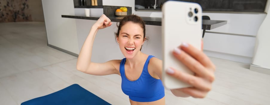 Young energetic girl does sports at home, shows her muscles on arm and takes selfie on smartphone, posing on fitness rubber mat in living room.