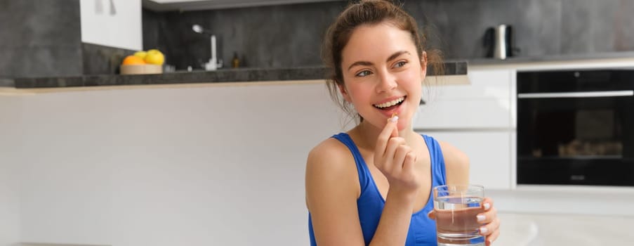 Healthy and happy fitness girl, taking vitamins, holding dietary supplement pill and glass of water, using buds after workout training session at home.