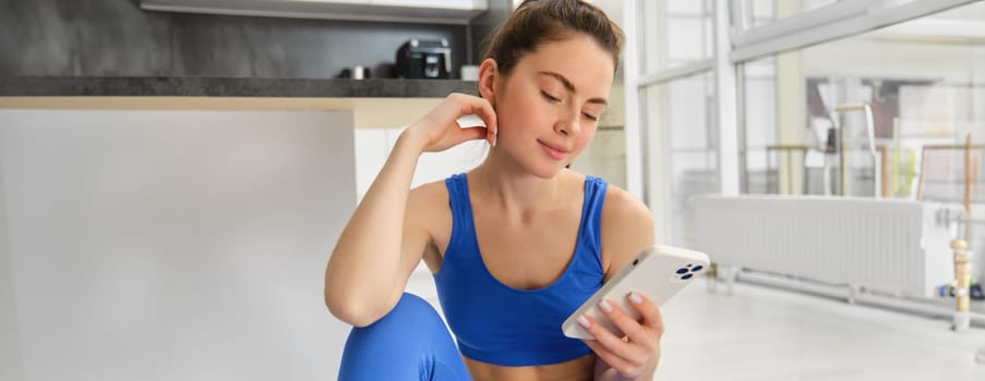 Young woman with smartphone does workout at home, using mobile phone app for sports training indoors, wears blue sportsbra and leggings.