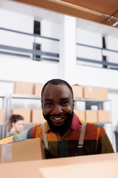 Smiling african american employee working at clients order, putting products in cardboard boxes, preparing products for shipping. Supervisor wearing industrial overall while carrying packages