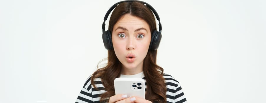 Portrait of happy young modern woman, female model in wireless headphones, smiling and looking at camera, holding smartphone, using mobile phone app, white background.