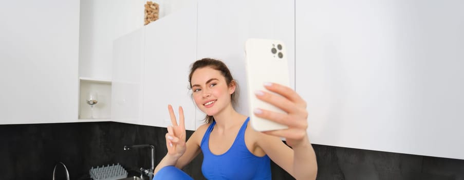 Portrait of beautiful woman, fitness blogger, taking selfie in sportsbra and leggings, posing in kitchen with smartphone.