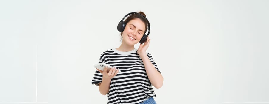 Young woman with smartphone listening to music, dancing to her favourite song in headphones, posing against white background.