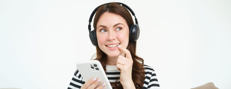 Image of girl in headphones, smiling while thinking, making decision, holding smartphone, making choice, online shopping, white background.