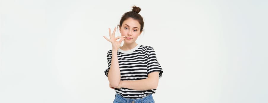 Portrait of brunette woman seals her mouth with promise not to tell anyone, zipping gesture, standing over white background.