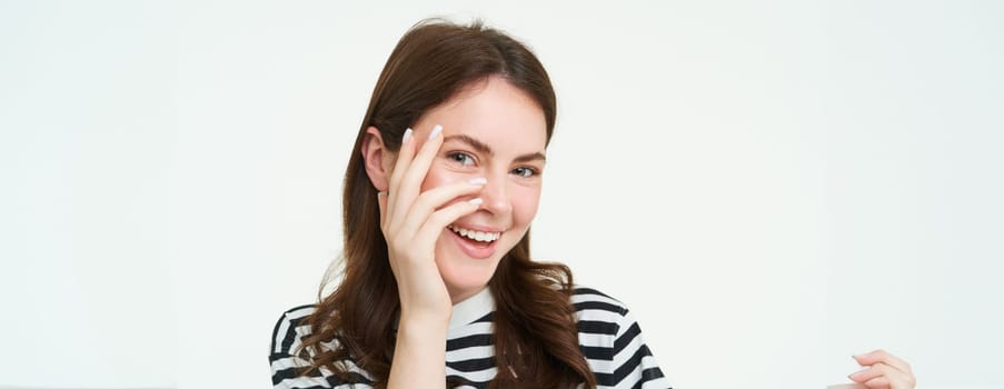 Portrait of attractive, smiling young woman, laughing and smiling, touches her face, standing over white background.