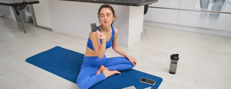 Image of smiling young woman, holding credit card, sitting on fitness mat with laptop, buying sport app subscription, workout video tutorials, doing exercises at home.