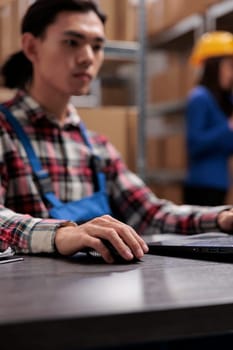 Young asian logistics manager analyzing goods supply on laptop. Warehouse worker doing inventory management on computer while sitting at desk with close up selective focus on hand