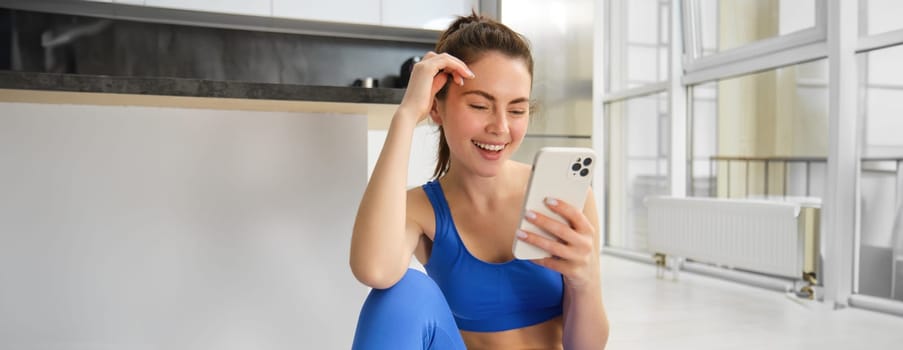 Image of young fit woman wearing sportbra and blue leggings, using smartphone app, does workout from home, sits on floor and follows online training video.