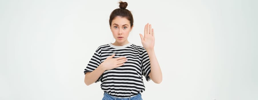 Image of woman makes promise, swears, raises one hand and puts palm on heart, being honest, stands over white background.