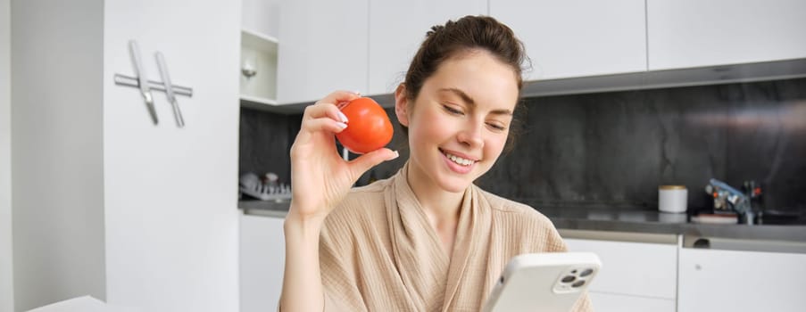 Image of smiling beautiful woman, sits in kitchen, holds smartphone and tomato, looks happy, orders groceries delivery to her home, using mobile phone app, buying online vegetables for her recipe.