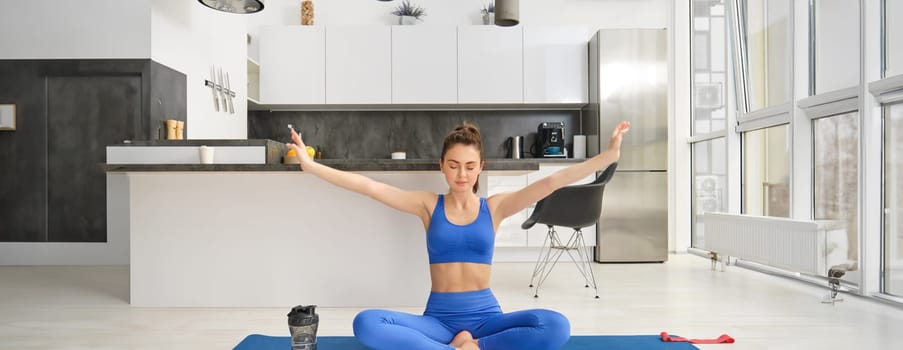 Young woman doing sports, meditating on yoga mat at home, holds hands sideways and breathing, relaxing mindful exercises.