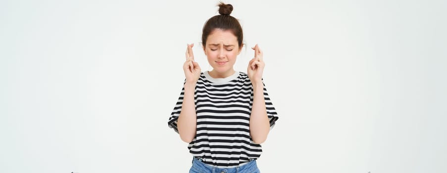 Portrait of hopeful young woman, makes wish, cross fingers for good luck, wishing, anticipating, standing over white background. Copy space