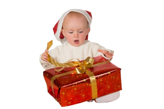 Excited cute little baby in a Santa hat untying a gold bow on a large red Christmas gift isolated on white