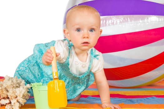 Conceptual studio image of a little baby with a bucket and spade and large striped beach ball crawling on a colourful towel at the seaside, on white with copyspace