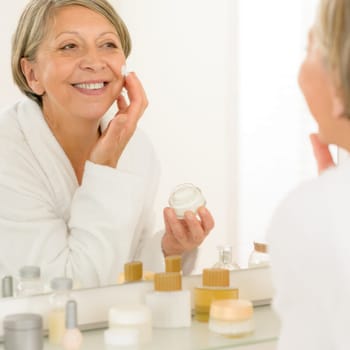 Smiling senior woman apply anti-wrinkles cream looking in bathroom mirror