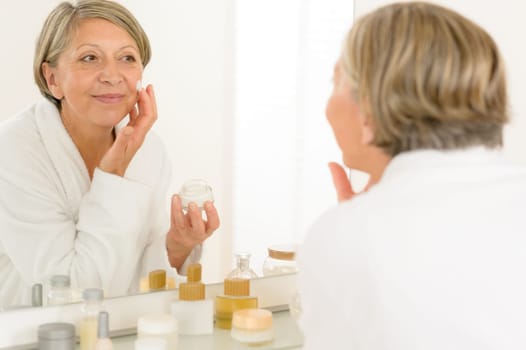 Senior woman looking in bathroom mirror applying anti-wrinkles face cream