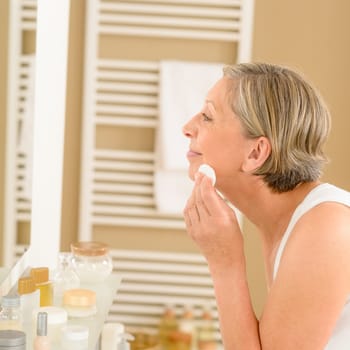 Senior woman clean face with cotton pad looking bathroom mirror