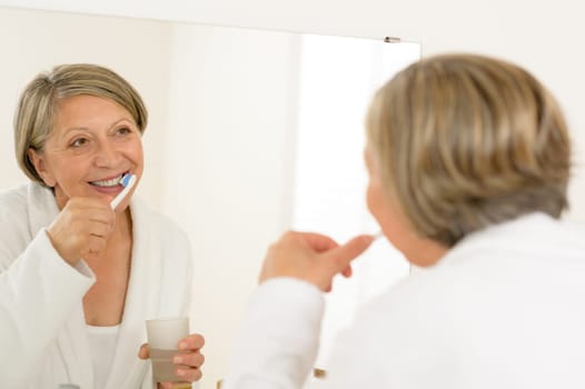 Mature woman brushing teeth with toothpaste looking in bathroom mirror