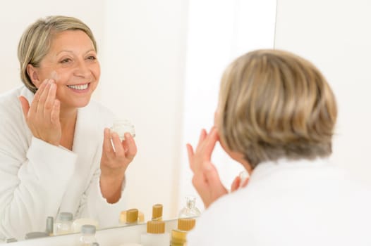 Senior woman looking in bathroom mirror and applying anti-wrinkles cream