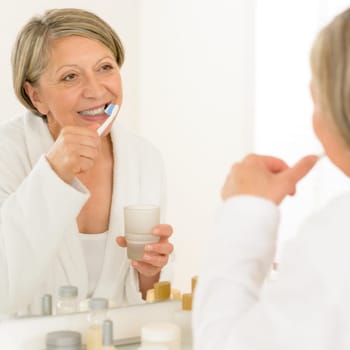 Senior woman brushing teeth looking at herself in bathroom mirror