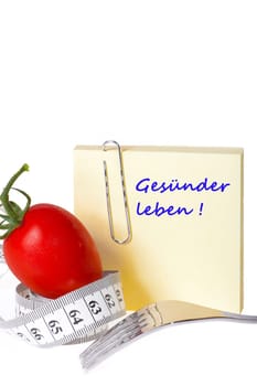 Sticky note with inscription: Healthier living - Yellow sticky note with the inscription: Healthier living with paper clip behind a tomato with measuring tape and a fork against a white background