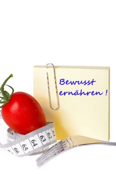 Sticky note with inscription: Eat consciously - Yellow notepad with the inscription: Eat consciously with paper clip behind a tomato with measuring tape and a fork against a white background