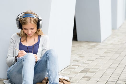 Student girl writing and listening to music leaning against wall