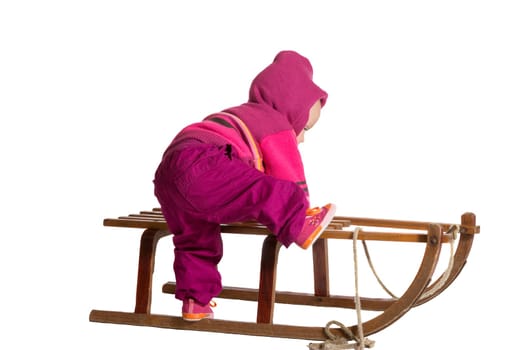 Rear view of a small toddler dressed in colourful winter woollies clambering onto a traditional wooden winter sled isolated on white