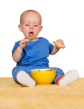 Cute little baby sitting on a rug with a yellow bowl between its legs eating biscuits with obvious pleasure and relish