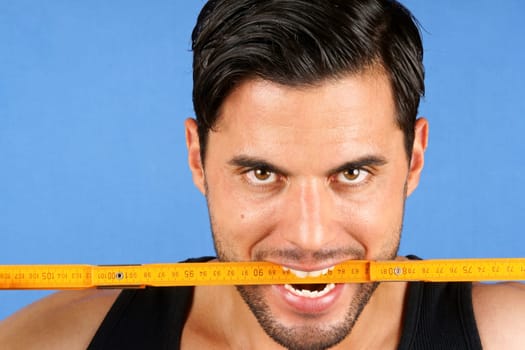 Angry handsome carpenter worker biting a wooden metric ruler over a blue background.