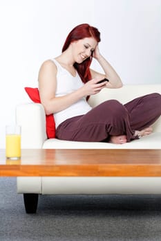 happy young woman sat on her white sofa at home smiling at text messages on her phone
