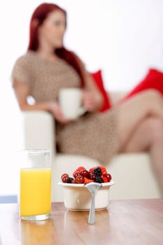 Bowl of fresh fruit and Orange juice on a coffee table with a young woman sat on a sofa in the background.