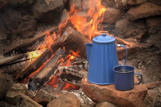 Enamel coffe pot percolator and blue enamel coffee cup resting on a rock near a campfire