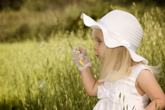 Little girl in a white dress and hat blowing soap bubbles in a field